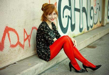 Photograph of a beautiful 20 year old redhead woman with shoulder-length hair in a loose bun. She is wearing a short, tight black print dress, red leggings, and black patent leather shoes with low heels. She is sitting near a wall covered with grafitti. She is looking at the viewer. She is smiling at the viewer with her mouth closed. Film photo, film grain, professional photo shoot.