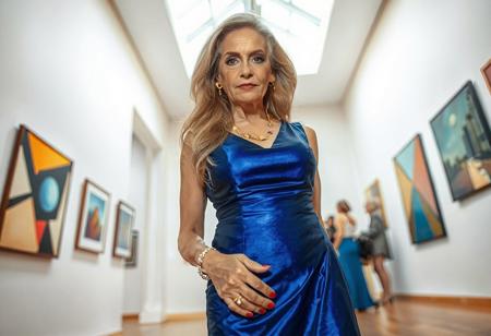 A well-built, mature woman of 45, with long blonde hair and tanned skin, is visiting a modern art gallery. She stands in front of the viewer, photographed in a full shot at a flat angle from below. She is wearing an elegant, shiny blue dress and blue high heels, along with golden and silver jewelry. Her face is made up with eye shadow and lipstick. She has perfect eyes and perfect hands with four fingers and one thumb, as well as red fingernails.  Her face is relaxed, and she is gazing directly into the viewer's eyes. Light flows through a white window in the ceiling.