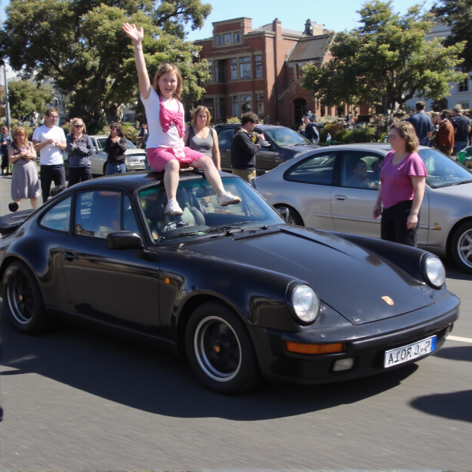 Porsche 911 GTS , london 1985, clear day.  the car is glossy black, There are people standing in the background, The girl from 1985 is sitting on the hood of a car, wearing few clothes. waving and greeting the passing car.  This photo was taken in 1985.   back to side camera angle , jpeg defects in photos <lora:Porsche 911:1>  <lora:amateurphoto:1>