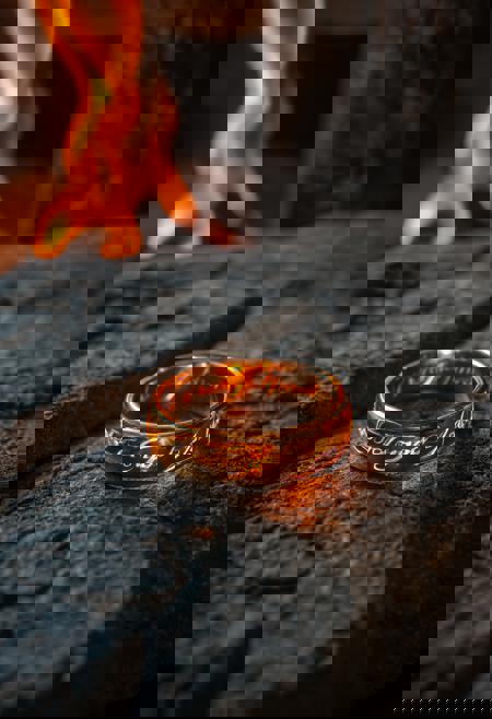 Hyper-realistic macro photograph of the One Ring from The Lord of the Rings, nestled within the heart of a roaring fireplace. The ring glows intensely, its golden surface reflecting the fiery orange flames that surround it. The elven inscriptions burn brightly, illuminated as if alive with ancient power. Embers and flickering sparks dance around, casting dynamic shadows on the rough stone hearth. The atmosphere feels alive and mesmerizing, with the ring radiating a dangerous, fiery allure amidst the crackling warmth of the fire