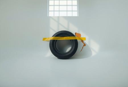 “Captured with Panavision Prime Cine optics: a minimalist studio scene showcasing a pastel white dome-shaped wall that curves smoothly into the floor, bathed in the soft glow of diffused cinematic lighting. Cube-shaped shadows, cast by a front-facing window grid, create depth and visual intrigue. At the precise center of the wall’s height, an old, weathered tyre rests, held in place by a broad strip of vibrant yellow duct tape. Beside the tyre, a jagged scar disrupts the wall’s otherwise smooth surface, introducing a raw, contrasting imperfection to the otherwise pristine setting. The premium optics highlight every texture— from the roughness of the tyre and wall to the bold contrast of the tape and scar, emphasizing the interplay of decay and order.”