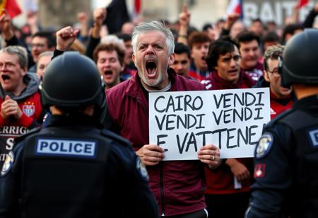 A photograph of a screaming, ragged, angry group of Torino FC fans, wearing a burgundy soccer vest, protesting with a poster in his hands that says "CAIRO VENDI E VATTENE!", police officers stand in the foreground with their backs to the viewer,