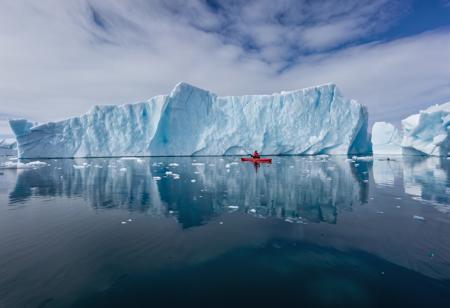 Raw photo, kayaking in Greenland: picture a lone kayaker amidst the icebergs of Greenland's icy waters. the photograph should contrast the deep blues and whites of the ice and water with the vibrant kayak, capturing the scale and isolation of the Arctic environment,