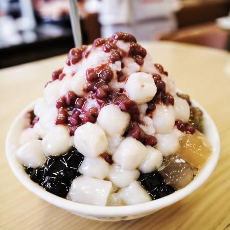 The image features a close-up of a bowl filled with a popular Taiwanese dessert often referred to as "boba tea ice" or "shaved ice." The bowl, which appears to be round and possibly white, contains a variety of toppings that may include cream, red beans, green beans, and possibly potato starch. A brown sauce is drizzled on top of the ice. The dessert is served with at least two spoons, suggesting it may be shared. The surroundings hint at the setting being a tea shop or a place where such desserts are commonly served, with other bowls in the background contributing to the atmosphere of the establishment. Despite the lack of text, the image seems to omit a casual and enjoyable vibe. , <lora:icetw:0.8>