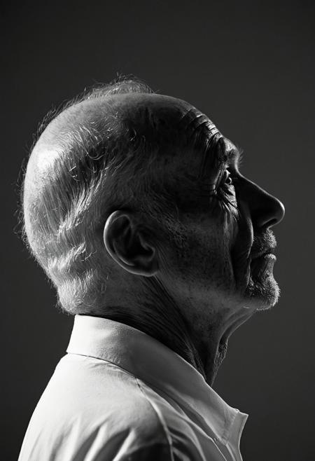 A striking black and white close-up portrait of an elderly man in profile against a dark, gradient background. The man wears a white garment and a black head covering, with his face turned to the right in a contemplative expression. His weathered skin and facial features are accentuated by dramatic side lighting. The shallow depth of field blurs the background while maintaining sharp focus on the man's distinctive features, creating a powerful portrait with documentary-style realism. Black and white portrait photography with dramatic side lighting and shallow depth of field.