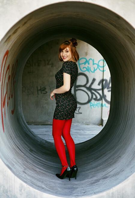 Photograph of a beautiful 20 year old redhead woman with shoulder-length hair in a loose bun. She is wearing a short, tight black print dress, red leggings, and black patent leather shoes with low heels. She is standing inside a large concrete storm culvert set into a concrete wall. The wall is covered with graffiti. Viewed from the side, her head turned toward the viewer. She is smiling at the viewer with her mouth closed. Film photo, film grain, professional photo shoot.