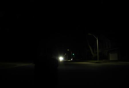 it is night. there is no light aside from the distant street light. the street is empty. the homes along the street are all dark. tall imposing trees stretch over the dark street. a man sits with his knees pulled up to his face and his arms wrapped around his legs. his face isn't visible but it seems he is sad. he sits in the street. I can not express enough how dark it is in this image and how upset I will be if it isn't dark.