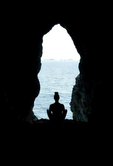 A coastal cave scene viewed from inside, looking outward toward the sea. The cave walls are dark, rugged, and textured, forming a natural frame with an uneven, jagged opening shaped like a distorted arch. Inside, a single person sits in silhouette on a rocky ledge, legs crossed, facing left. Their back is upright, head slightly tilted up in a contemplative pose, and hair tied in a bun. No facial features are visible due to strong backlighting.
Outside the cave, the sea glows under daylight—calm, silver-blue water stretching to the horizon with faint landmasses in the distance. The sky shifts from bright blue to pale near the horizon. The contrast between the cave’s shadowy interior and the glowing outside world creates a dramatic silhouette effect.
Use a naturalistic palette: dark browns and charcoal for the cave, soft blues and silvers for the sea and sky. Lighting is high-contrast; rim light gently outlines the cave edges and the seated figure. The mood is tranquil and introspective, evoking awe in nature’s scale and serenity.