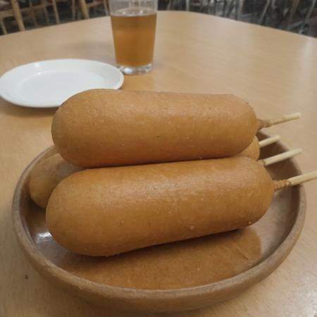 A photorealistic image of a delicious 9hotdog placed on a plate in a restaurant setting. The 9hotdog should be golden brown, with a crispy exterior and a juicy interior visible at one end where it's bitten off. The plate can be white or a light color to contrast with the golden hue of the 9hotdog. There might be a small dish of mustard or ketchup on the side for dipping. The table should have a clean tablecloth, and the lighting should be warm, highlighting the appetizing look of the 9hotdog. The background can have subtle hints of a restaurant ambiance, like a glass of water or some cutlery, <lora:cornhotdog:0.65>, (bottle of soda),<lora:applesoda:0.75>