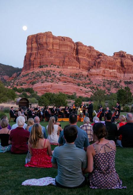 couples on a large lawn in front of a wall of natural red rock formation, listening to an orchestra on a warm summer night in the foothills, half moon, sitting resting on hands, some snuggling, one couple kissing,  conductor smiling, close up on couples