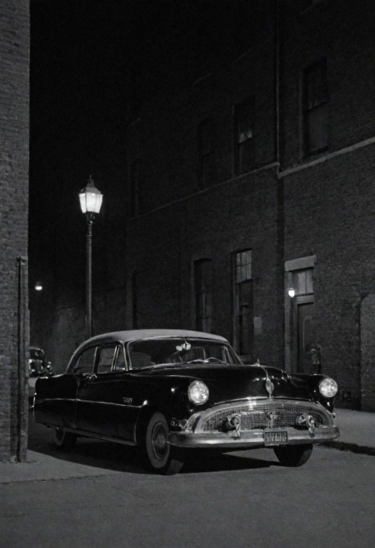 A classic black car parked on a street at night, near a building and a light pole.