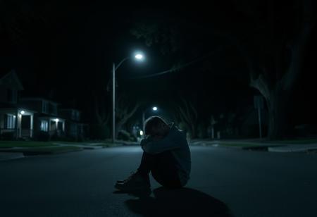 it is night. there is no light aside from the distant street light. the street is empty. the homes along the street are all dark. tall imposing trees stretch over the dark street. a man sits with his knees pulled up to his face and his arms wrapped around his legs. his face isn't visible but it seems he is sad. he sits in the street. I can not express enough how dark it is in this image and how upset I will be if it isn't dark.