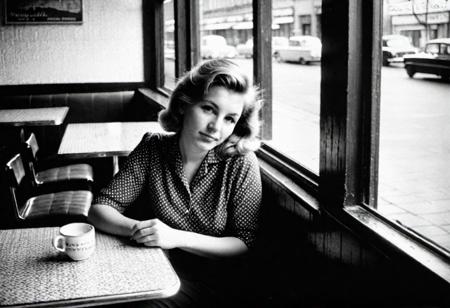 german woman 30 years old sitting in a cafe in east berlin in 1962, watching outside, face in shadow, low light, full body shot from side, 1960 soviet look, dark mood, ilford hp5 plus 400, medium format camera,artsy, shadows, wide angle, contact test, damaged print, faded, grainy,blurry, pixelated, motion blur, vignette, high contrast