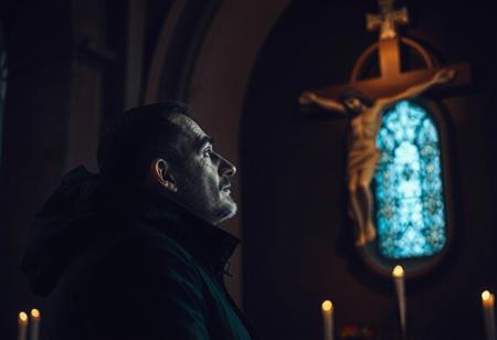 professional photo, side view photo of Caucasian man wearing a dark blue parka with dark features and a serious contemplative expression looking up at a plaster statue of jesus on the cross mounted on the wall of a church, the man and Jesus are shown in profile, the scene is very dark and underexposed with only the man's and Jesus' face lit by warm, dim candle light, everything else is dark shadows