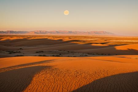 A desolate desert landscape with rolling sand dunes and distant mountains, bathed in the golden glow of an orange sun setting behind one mountain peak. The sky is cloudless, creating long shadows on the ground, and there's a hint of mist or fog around. In the distance, another moon can be seen.
  <lora:MJ52:1>