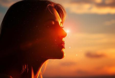 Softly fading sunlight casts a warm glow on the woman's serene face, her silhouette a celestial canvas of swirling galaxies and shimmering stars. Ethereal light dancing across her features, as a radiant aura envelops her, merging harmoniously with the sky's gentle hues. The cosmos within her outline twinkles like diamonds scattered across the heavens, taking refuge within her essence.
