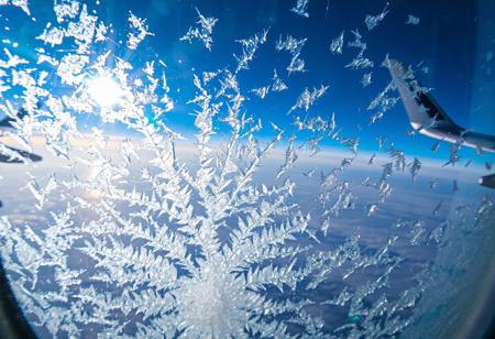 JNANX,luminous white silhouette,cyan glow,emerald highlights,flickering outline,dreamlike movement,
"A close-up view from an airplane window reveals intricate ice crystals forming on the outer pane. The blue sky and a portion of the aircraft's wing are visible in the background, distorted slightly by the frosty accumulation. Numerous delicate, feathery ice patterns spread across the window, with a denser, more concentrated cluster at the bottom center, almost appearing to grow upwards. Sunlight reflects off the icy surface, creating subtle glints. The overall impression is one of cold beauty, showcasing the unique atmospheric conditions encountered at high altitudes. The textures of the ice are varied, from fine dust-like particles to more defined, branching structures, suggesting a dynamic freezing process. The window itself has some minor smudges or imperfections, adding to the authenticity of the aerial perspective."