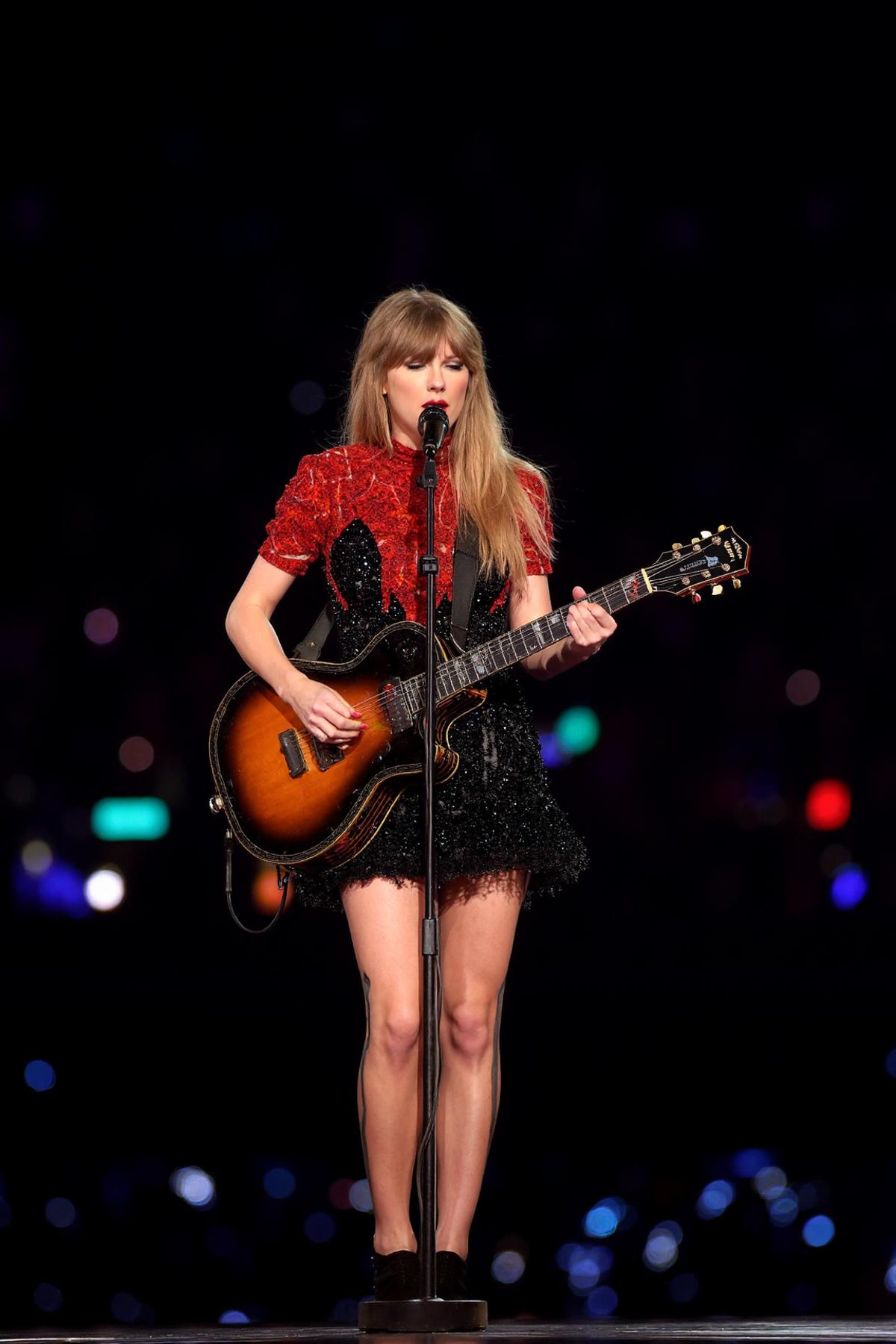 A full-body photograph of taySwift playing guitar while singing into a microphone, wearing a striking black and red dress that catches the light, against a dark black background. she holds a microphone in his other hand, posing with a sense of charisma and stage presence. on stage and lit by colored lights with fans cheering in the background