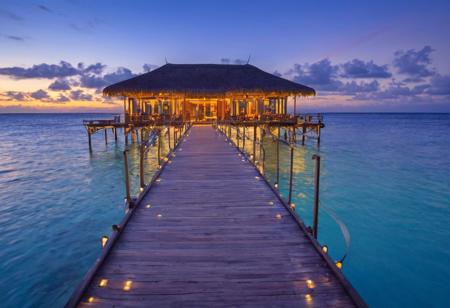 overwater bungalow, sunset, ocean view, tropical, luxury resort, vivid colors, high dynamic range, architectural lighting, modern design, wooden walkway, reflection on water, blue hour, clear sky, holiday destination, LED lights, Christmas tree visible, serene, high-resolution image, tranquil scene, travel photography.