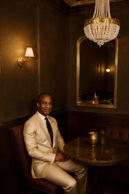 RAW Photo, photo of light-skinned African American man sc_chad3 <lora:sc_chad3-06:1> sitting introspectively in a booth in a Parisian-style cafe, contemplative smile, wearing tailored (tom ford suit) with ivory dress shirt and silk burgundy gucci tie with gold accents, deep mahogany walls, ornate gold-framed mirrors, ambient glow, dim interior, soft daylight filtering through lace curtains, aged wooden floor, brass espresso machine, porcelain, shadowed corners, serene atmosphere, nostalgic atmosphere, (vanity fair magazine), (photo by Sebastião Salgado)