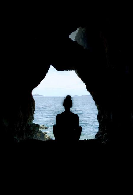 A coastal cave scene viewed from inside, looking outward toward the sea. The cave walls are dark, rugged, and textured, forming a natural frame with an uneven, jagged opening shaped like a distorted arch. Inside, a single person sits in silhouette on a rocky ledge, legs crossed, facing left. Their back is upright, head slightly tilted up in a contemplative pose, and hair tied in a bun. No facial features are visible due to strong backlighting.
Outside the cave, the sea glows under daylight—calm, silver-blue water stretching to the horizon with faint landmasses in the distance. The sky shifts from bright blue to pale near the horizon. The contrast between the cave’s shadowy interior and the glowing outside world creates a dramatic silhouette effect.
Use a naturalistic palette: dark browns and charcoal for the cave, soft blues and silvers for the sea and sky. Lighting is high-contrast; rim light gently outlines the cave edges and the seated figure. The mood is tranquil and introspective, evoking awe in nature’s scale and serenity.
