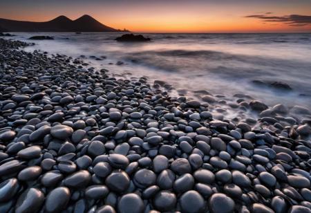 The photo presents a dramatic and dynamic coastal landscape at sunset. In the foreground is a collection of dark volcanic round pebbles with soft, flowing water pouring over them, creating a milky and gentle texture through a long exposure. The sun setting on the horizon colors the sky in vivid shades of orange and blue, creating a strong contrast to the dark silhouette of the coastline.