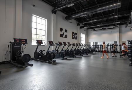 A gym with numerous mills, individuals exercising with dumbbells, a window, pristine walls, gray epoxy resin, and a dark open ceiling, featuring typography.