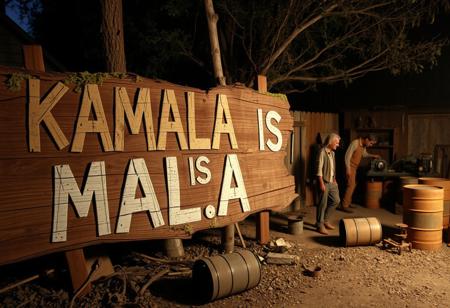 Create an image of a large, decaying wooden sign that reads “KAMALA IS MALA” in faded, roughly painted letters. The sign appears weathered, with mold, moss, and splintered wood giving it a neglected, ominous feel. In the background, set up a chaotic, makeshift backyard resembling a rundown meth lab. A few rugged redneck characters, dressed in worn-out, dirty clothes, are busy with rusty equipment and improvised setups. The area is cluttered with scattered debris, oil barrels, and old machinery. The lighting is dim and gritty, casting heavy shadows to emphasize the grungy, disorganized environment.