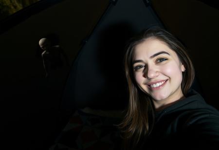 A woman taking a selfie inside a small modern tent at night. She is smiling for the camera and winking. Behind her is the wall of the dark tent illuminated by the camera flash. In the top left of the image the sheer and partially see-through mesh of the tent is visible. A scary disturbing eerie creature of some sort is barely visible through the mesh in the top left of the image and is leaning over the outside of tent. The woman taking the selfie is oblivious to its presence since it is outside and not easily visible in the image. The creature is tall and strange and very dimly lit by moonlight but is primarily visible via its silhouette.