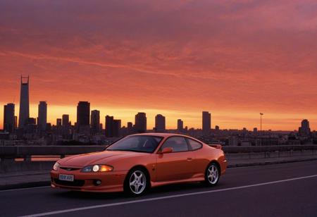 (1996 Hyundai Tiburon RD) motor vehicle, no humans, city, sky, outdoors, scenery, orange sky, road, building, cityscape, sunset, vehicle focus, skyscraper, sports car, shadow, gradient sky, red sky, evening, cloud, street, skyline, twilight, spoiler \(automobile\), orange background, dusk, red background, water, solo, bridge,
