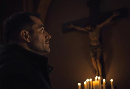 professional photo, side view photo of Caucasian man wearing a dark blue parka with dark features and a serious contemplative expression looking up at a plaster statue of Jesus on the cross mounted on the wall of a church, the man and Jesus are shown in profile, the scene is very dark and underexposed with only the man's and Jesus' face lit by warm, dim candle light, everything else is dark shadows