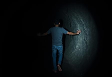 a very dark dim barely visible image at night of a man walks down a midnight black tunnel carved into the earth. there is no light, no daylight, and he is only barely visible. he faces away from the viewer as he walks deeper into the earth. his arms are stretched out slightly, timidly, as if he is afraid of what he might touch in the darkness that surrounds him. He is wearing a grey t-shirt and blue jeans. he is barefoot. the dark tunnel is wet and drips of moisture can barely be seen falling from the craggy surface of the rock. the tunnel seems endless in its depth and darkness. the image is one of nearly complete darkness.