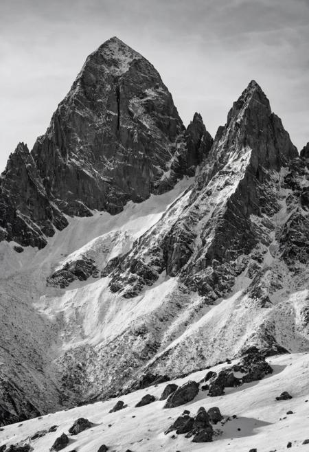 A minimalist black and white landscape of towering mountain peaks dusted with snow under a clear, bright sky. The rugged textures and stark contrasts between the black rock and white snow create a dramatic yet simple composition. The color grading should bring out the deep blacks and crisp whites, emphasizing the sharp contrasts and clean lines of the landscape. Shot on a Canon EOS 5D Mark IV with a Canon EF 24-70mm f/2.8L II USM lens by Marta Bevacqua.