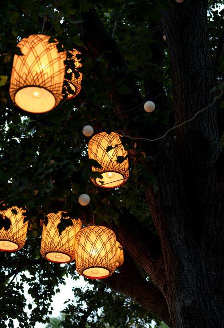 A low-angle shot captures a cluster of illuminated, geometric paper lanterns hanging from the sturdy branches of a mature tree. The lanterns are primarily cylindrical with rounded tops and bottoms, featuring a repeating pattern of radiating lines in warm orange and white hues. The light emanating from within casts a soft glow on their surfaces, highlighting the texture of the paper and the intricate design. Some lanterns appear partially obscured by the dense green foliage of the tree, creating a sense of depth and layering within the frame. The leaves are varied in shape and size, suggesting different types of foliage intertwined. A few smaller, spherical white lights are also visible, strung along a dark cord that weaves through the branches alongside the lanterns. The bark of the tree is rough and textured, with visible knots and variations in color. The overall lighting suggests either dusk or dawn, with a soft, natural light filtering through the leaves and illuminating the scene. The focus is sharpest on the central lanterns, gradually softening towards the edges of the frame, drawing the viewer's eye to the illuminated focal point. The scene evokes a feeling of warmth, tranquility, and perhaps a festive or decorative outdoor setting.