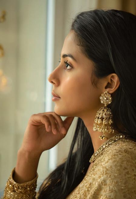 A professional fashion portrait captures a young South Asian woman in traditional golden attire, photographed in profile against a softly lit interior background. The woman wears ornate golden chandelier earrings and an embellished golden dupatta draped across her shoulders. Natural side lighting from a window creates a warm, ethereal atmosphere in the room. The woman's dark hair is styled sleek and straight, framing her refined facial features in profile view. Her hands are positioned gracefully beneath her chin in a contemplative pose. The composition utilizes shallow depth of field, maintaining sharp focus on the subject while the background remains softly blurred. Professional portrait photography with soft natural lighting, shallow depth of field, and precise focus on subject against blurred background.
