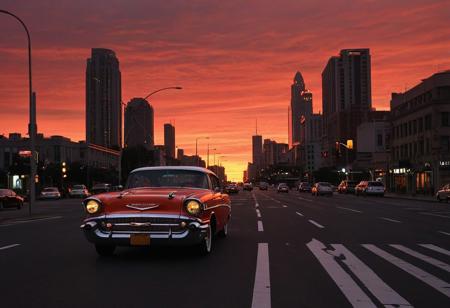 car, motor vehicle, no humans, road, building, city, lamppost, sky, outdoors, scenery, orange sky, street, sunset, cityscape, vehicle focus, skyscraper, red sky, evening, gradient sky, shadow, traffic light, twilight,  full-size car, cloud, dusk, red background, crosswalk, window, skyline, road sign, (1957 Chevrolet Bel Air)