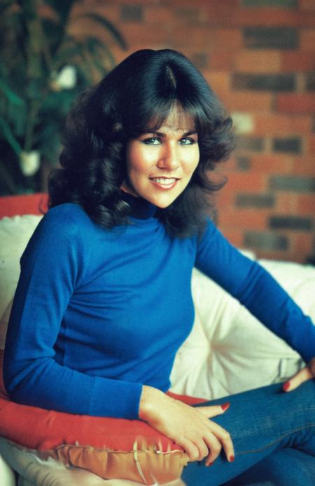 A (sfw:99.8) professional photo of lindalusardi woman at home in her apartment, smiling at viewer, surrounded by (1980s furniture:1.4), for a women's interest magazine, wearing a tight but concealing red (turtleneck:1.2) sweater and long dark blue jeans, large breasts under sweater, (best quality:1.2), (intricate details)