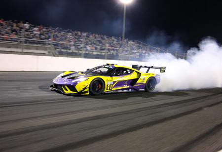 "This image portrays a race car in full motion on a track, with a cloud of smoke trailing behind it. The car's striking yellow and purple color scheme, along with black accents, creates a visually captivating appearance. The smoke adds a sense of speed and intensity to the scene. The well-lit track and the enthusiastic crowd in the stands contribute to the excitement and intensity of the race."