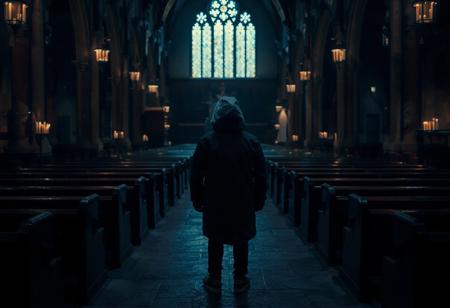 A small lone figure in a dark blue parka with the hood pulled up stands in the middle of an old cathedral dimly lit by candles, twilight, very dark and underexposed, pews line the walls of the church in the traditional layout, overhead view