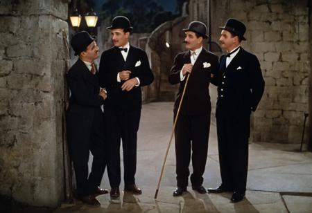 1931 Charlie Chaplin City Lights, color image depicts a classic scene with three men on a sidewalk at night. The layout is horizontal, with the subjects positioned in the foreground. On the left, a man with a mustache, wearing a bowler hat, a suit, and holding a cane, is tipping his hat. He has light skin and a distinctive expression. Next to him, another man, Charlie Chaplin, Chaplin's Tramp, also with light skin, dressed in a tuxedo and top hat, is leaning slightly and smiling. Both men are interacting with a uniformed officer standing on the right, who has his hands on his hips and is holding a baton. The background features a stone wall and steps, with a lamppost providing illumination. The overall atmosphere is reminiscent of early 20th-century cinema. 1930s movies style