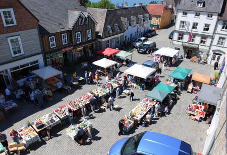 small town Saturday market day, sunny, northerly, July, crisp, near the sea,, village square filled with 40 tables, 20 booths, crafts, homemade food, carvings, buyers, sellers, colorful, chocolate sop, bakery, parked cars, produce, berries, pottery, wool weaving, hot dog grilling, bookstore, bank building, tea room, view from above