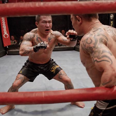 An MMA fighter in the midst of a battle inside the Octagon cage. He's in a dynamic fighting stance, muscles tensed, eyes focused on his opponent. The background shows the cage, referee, and a blurred crowd. The scene should be as realistic as a high-resolution photograph, capturing the intensity, sweat, and determination of a live MMA match. <lora:gan2_video:0.75>,ganjo