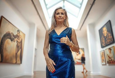A well-built, mature woman of 45, with long blonde hair and tanned skin, is visiting a modern art gallery. She stands in front of the viewer, photographed in a full shot at a flat angle from below. She is wearing an elegant, shiny blue dress and blue high heels, along with golden and silver jewelry. Her face is made up with eye shadow and lipstick. She has perfect eyes and perfect hands with four fingers and one thumb, as well as red fingernails.  Her face is relaxed, and she is gazing directly into the viewer's eyes. Light flows through a white window in the ceiling.