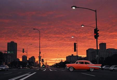 car, motor vehicle, no humans, road, building, city, lamppost, sky, outdoors, scenery, orange sky, street, sunset, cityscape, vehicle focus, skyscraper, red sky, evening, gradient sky, shadow, traffic light, twilight,  full-size car, cloud, dusk, red background, crosswalk, window, skyline, road sign, (1957 Chevrolet Bel Air)