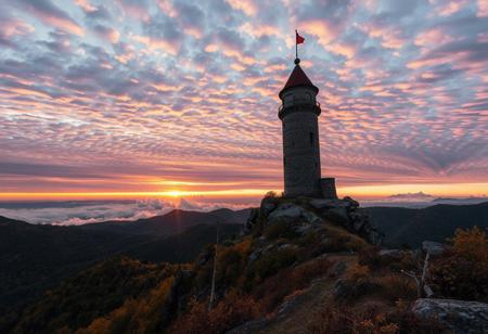 A tower of solitude in the mountains, a dramatic sunset sky filled with layered clouds, surrounded by autumn vegetation and rocky hills. Epic photography by Evgeni Dinev.