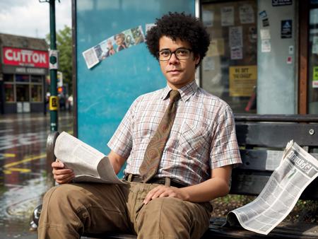 Richard Ayoade as Moss, parted afro, plaid_shirt, paisley tie, tweed_pants,, portrait stock photo middle-aged man in at a bus stop, sitting on bench, scruffy beard, messy hair, t-shirt, covering head with newspaper, London street corner, rainy day, (masterpiece:1.2) (photorealistic:1.2) (bokeh) (best quality) (detailed skin) (intricate) (8k) (HDR) (cinematic lighting) (sharp focus) <lora:Richard-Ayoade-v10:0.7>