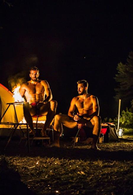 Two hairy chested fit italian men in a campsite, shirtless, sitting in a camping tend, illuminated by a fire lit in front of the tent, in a dark night