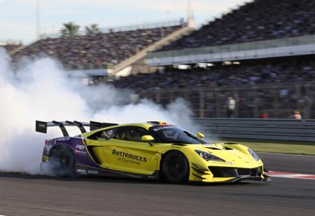 "This image portrays a race car in full motion on a track, with a cloud of smoke trailing behind it. The car's striking yellow and purple color scheme, along with black accents, creates a visually captivating appearance. The smoke adds a sense of speed and intensity to the scene. The well-lit track and the enthusiastic crowd in the stands contribute to the excitement and intensity of the race."