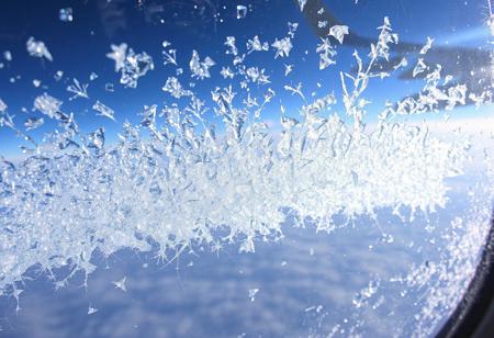 JNANX,luminous white silhouette,cyan glow,emerald highlights,flickering outline,dreamlike movement,
"A close-up view from an airplane window reveals intricate ice crystals forming on the outer pane. The blue sky and a portion of the aircraft's wing are visible in the background, distorted slightly by the frosty accumulation. Numerous delicate, feathery ice patterns spread across the window, with a denser, more concentrated cluster at the bottom center, almost appearing to grow upwards. Sunlight reflects off the icy surface, creating subtle glints. The overall impression is one of cold beauty, showcasing the unique atmospheric conditions encountered at high altitudes. The textures of the ice are varied, from fine dust-like particles to more defined, branching structures, suggesting a dynamic freezing process. The window itself has some minor smudges or imperfections, adding to the authenticity of the aerial perspective."