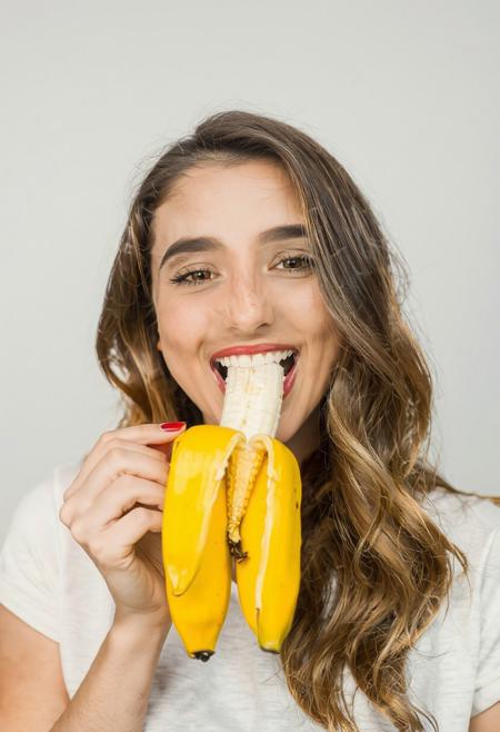 beautiful woman eating a half-peeled banana, mouth closed around banana stalk, banana held in her hand, banana peel moved down to midway, woman delighted.